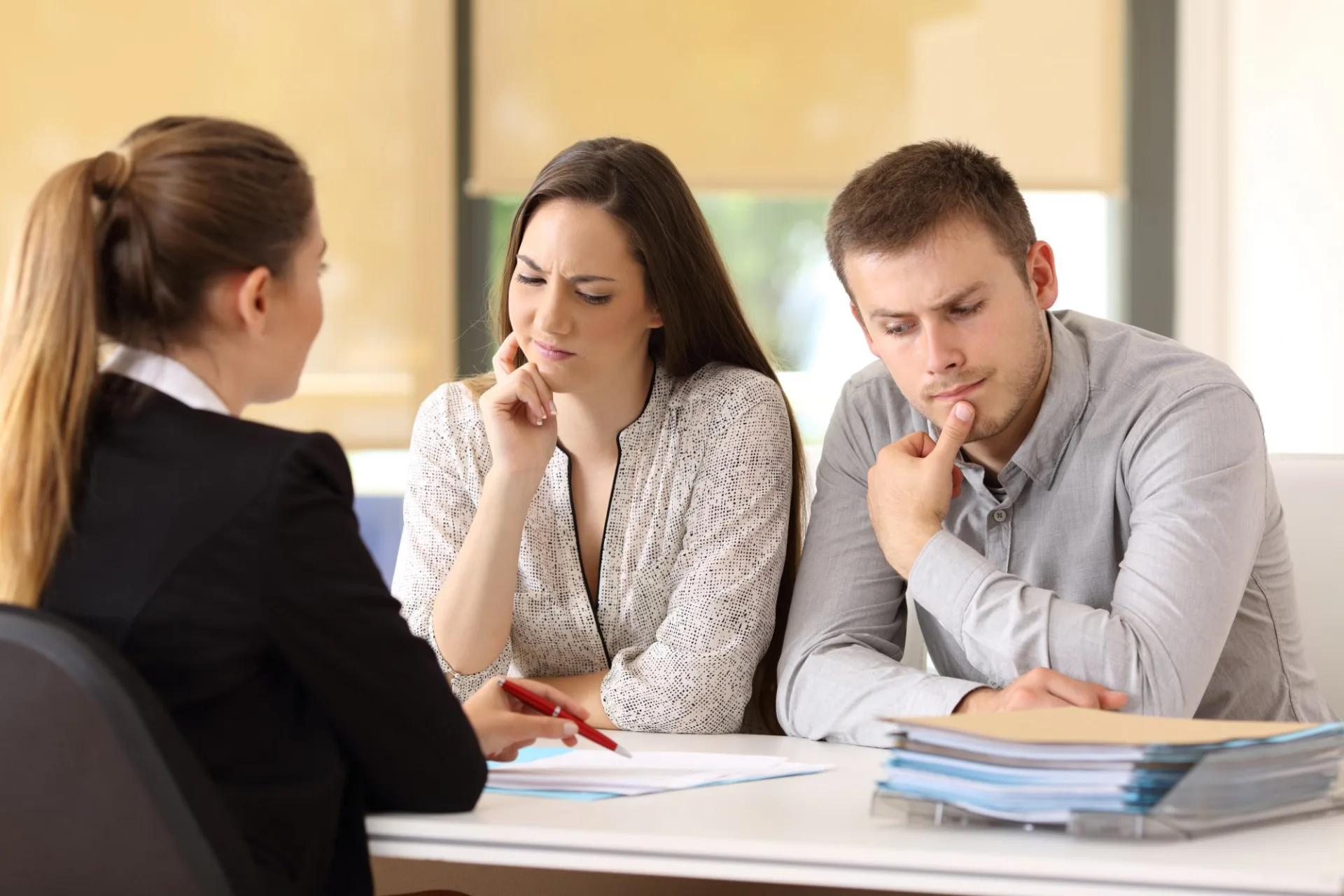 A couple sits at a desk looking concerned while meeting with a professional who is holding a pen and pointing to paperwork.