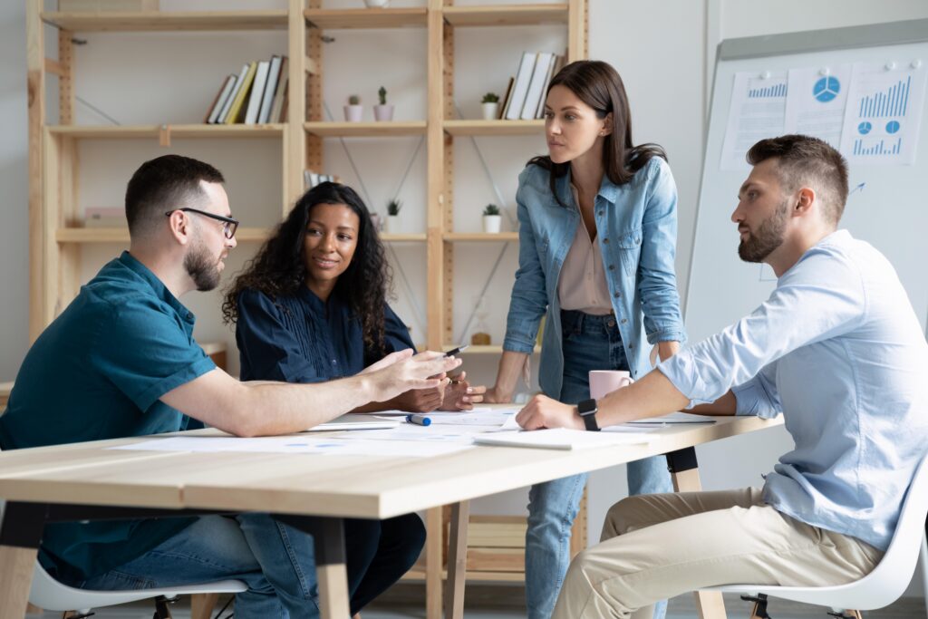 Four coworkers in a bright office discuss charts and reports around a wooden table, with a whiteboard of graphs in the background.